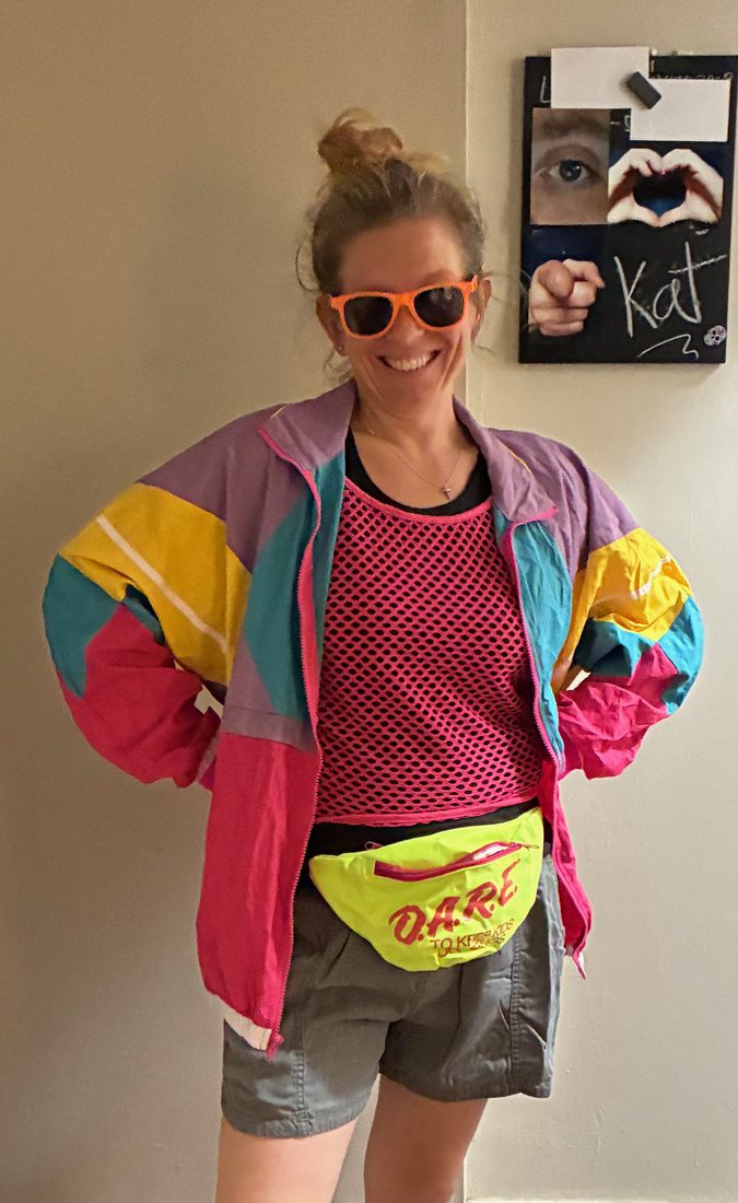 Kat smiling in orange sunglasses, a color-block windbreaker, and a neon fanny pack